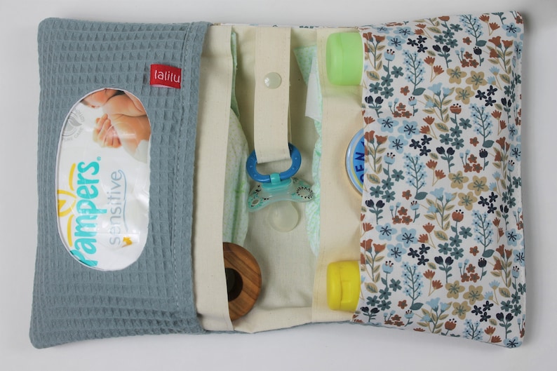 May include: A blue and white diaper bag with a floral pattern. The bag has a zippered closure and a flap with a snap closure. The bag is open and shows the inside, which is lined with a cream-colored fabric. The bag contains a variety of baby items, including diapers, wipes, a pacifier, and a bottle of lotion. The bag also has a small pocket on the outside for a phone or keys.