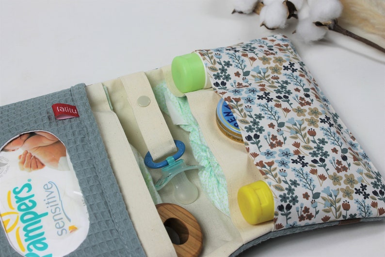 May include: A light blue diaper bag with a floral pattern on the outside. The bag is open and shows the inside, which has several pockets and compartments. The bag is filled with baby supplies, including diapers, wipes, a pacifier, a bottle of lotion, and a tube of diaper rash cream. The bag also has a small tag that says "lalilu".