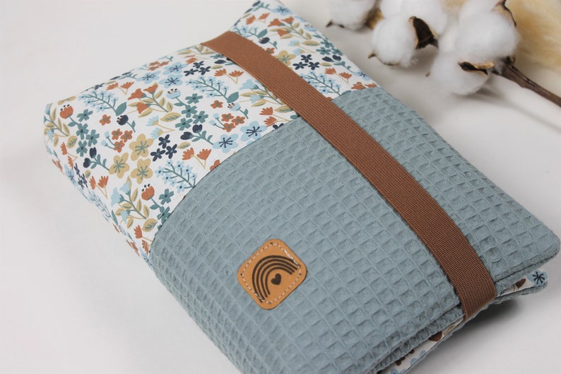 May include: A blue and white floral fabric pouch with a brown strap and a leather patch with a rainbow and heart design. The pouch is made of a soft, textured fabric.
