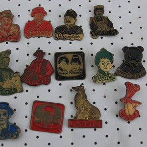 Peut inclure: Une collection de 13 épingles en émail vintage représentant divers personnages et logos. Les épingles sont principalement rouges, vertes et noires avec des accents dorés. Parmi les personnages, on trouve un ours en peluche, un chien et un chevalier. Les logos incluent "Kladsie", "Ivanhoe", "Lassie" et "Swartjie".