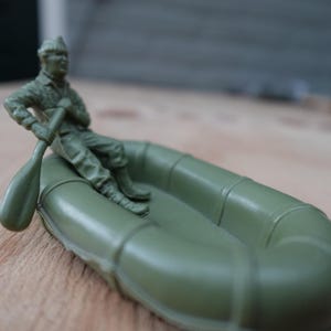 Può includere: Una figura di soldato giocattolo di plastica verde seduta su una barca gonfiabile verde, che tiene un remo. Il giocattolo è su una superficie di legno. La barca è un giocattolo classico, spesso associato a temi militari o di avventura.