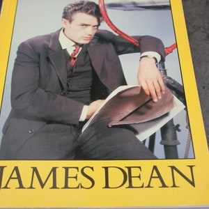 Può includere: Una copertina di libro gialla con il titolo "JAMES DEAN" in testo nero. La copertina presenta una foto in bianco e nero di James Dean con un abito e una cravatta, seduto su una struttura metallica.