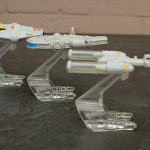Op de afbeelding: Drie Star Wars ruimteschip speelgoedmodellen op heldere plastic displays. De ruimteschepen zijn wit met gele accenten. De modellen zijn in een rij van links naar rechts gerangschikt.