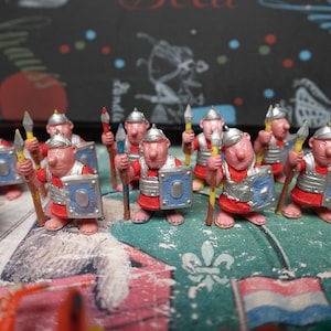 Op de afbeelding: Een verzameling Romeinse speelgoedsoldaten. Elke figuur draagt een rood en zilveren harnas, met een speer en schild. De schilden zijn blauw met een zilveren rand. De figuren staan op een kleurrijk oppervlak.