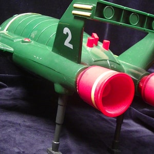 Puede incluir: Una nave espacial de juguete verde y roja con el número "2" en el lateral. La nave espacial tiene dos puertos de escape rojos y tren de aterrizaje.