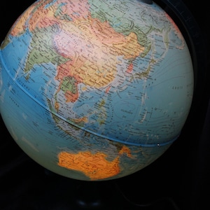 Op de afbeelding: Een blauwe globe met een gedetailleerde wereldkaart, waarop continenten, oceanen en landen te zien zijn. De globe wordt van de zijkant belicht, waardoor de continenten in oranje en geel worden geaccentueerd.