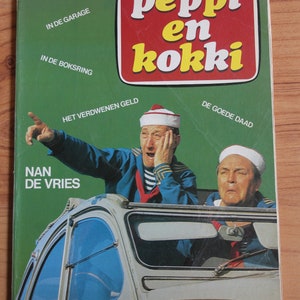 Op de afbeelding: Een groene en rode comicboekomslag met de titel "peppi en kokki" en het nummer "2" in een cirkel. De omslag toont twee mannen in een auto, een wijst en de ander kijkt verbaasd. De tekst op de omslag omvat "IN DE GARAGE", "IN DE BOKSRING", "HET VERDWENEN GELD", "DE GOEDE DAAD", "NAN DE VRIES".