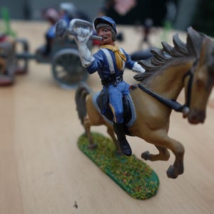 Puede incluir: Un soldado de juguete a caballo, tocando una trompeta. La figura lleva un uniforme azul con una bufanda amarilla. El caballo es marrón con una brida negra. Otros soldados de juguete y un carro están en el fondo.