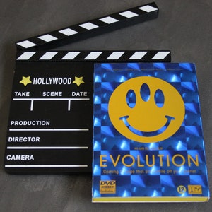 Può includere: Una clapperboard nera e bianca con la scritta "HOLLYWOOD" stampata su di essa. Una custodia per DVD blu con una faccia sorridente gialla e il testo "EVOLUTION" è accanto alla clapperboard.
