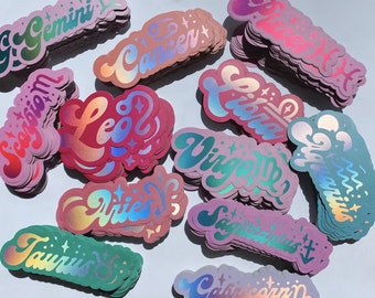 May include: A collection of holographic stickers featuring the astrological signs of the zodiac. The stickers are in various colors, including pink, blue, green, and purple. Each sticker features a different zodiac sign, including Gemini, Cancer, Leo, Virgo, Libra, Sagittarius, Capricorn, Taurus, Pisces, and Aquarius.