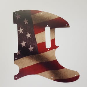 May include: Guitar pickguard with a design of the American flag. The pickguard has a distressed appearance with red and white stripes and white stars on a blue field. It is shaped with several screw holes.