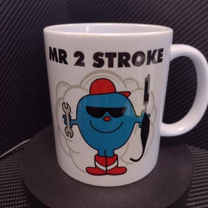 Può includere: Una tazza in ceramica bianca con un personaggio dei cartoni animati blu che tiene una chiave inglese e uno strumento nero. La tazza ha la scritta "MR 2 STROKE" stampata su di essa.