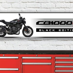 Op de afbeelding: Een zwarte motorfiets met een witte achtergrond. De motorfiets heeft een zwart zadel en een zilveren uitlaatpijp. De tekst "CB1000R BLACK EDITION" wordt weergegeven op de banner.