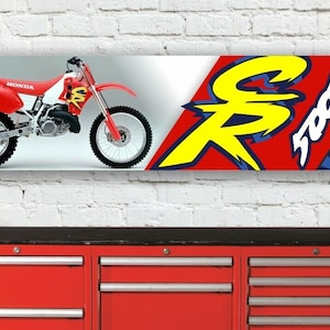 May include: A banner featuring a red and white Honda CR500R dirt bike. The banner has the Honda logo and the text "CR 500R" in yellow and white on a red background. The banner is hanging on a white brick wall.