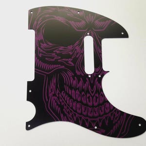 May include: Black guitar pickguard featuring a purple skull design. The pickguard has cut-outs for pickups and controls. The skull design is detailed and covers most of the pickguard's surface.