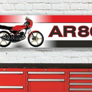 Può includere: Una motocicletta Kawasaki AR80 rossa e nera su sfondo bianco. La motocicletta ha una sella nera e uno scarico nero. La motocicletta è su uno sfondo bianco con il testo "AR80" in lettere nere.