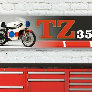 Könnte beinhalten: Ein schwarz-weißes Banner mit einem roten und weißen Streifen am unteren Rand. Das Banner zeigt ein weißes Yamaha TZ 350 Motorrad mit blauen und roten Akzenten. Der Text "TZ 350" ist in großen roten Buchstaben mit goldener Umrandung.