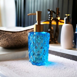 May include: A blue glass soap dispenser with a gold pump and a geometric pattern. The dispenser is sitting on a white surface.