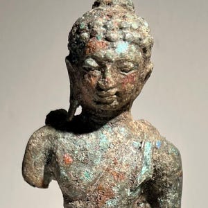 Puede incluir: Una estatua de bronce desgastada de una figura de pie, posiblemente una deidad. La estatua tiene una expresión serena y rasgos faciales detallados. El bronce tiene una pátina verde y marrón, con toques de azul y rojo.