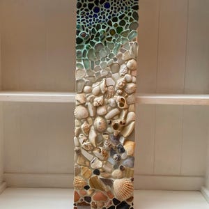 May include: A rectangular mosaic art piece featuring a gradient design. The top section has dark blue and green glass pieces, transitioning to clear and white shells and stones. The bottom section is composed of various seashells and pebbles.