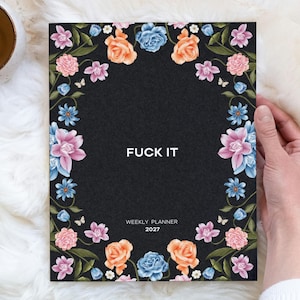 May include: A black weekly planner for 2027 featuring a floral border with pink, orange, and blue flowers and green leaves. The text FUCK IT is printed in white in the center, with WEEKLY PLANNER 2027 printed in smaller white text below.