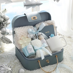 Könnte beinhalten: Eine blaue Geschenkbox gefüllt mit einem weißen Becher, einer Kerze, einem Notizbuch und einer flauschigen Decke, gebunden mit hellblauen Bändern. Die Box steht auf einer weißen, flauschigen Oberfläche. Im Hintergrund ist ein kleiner Weihnachtsbaum zu sehen.