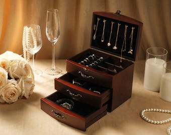 May include: A wooden jewelry box with three drawers and a hinged top, open to reveal necklaces. The box is a rich brown color with ornate drawer pulls. The drawers are lined with black velvet. Champagne glasses, candles, and a bouquet of roses are in the background.
