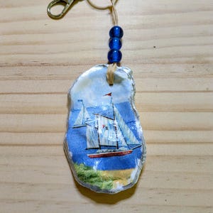 May include: A keyring featuring a natural oyster shell painted with a sailboat scene on a blue background. The shell is attached to a silver metal keyring and lobster clasp by a tan cord adorned with three small, round, translucent blue beads.