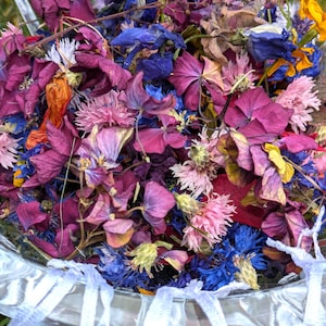Op de afbeelding: Een heldere glazen kom gevuld met een mengsel van gedroogde bloemblaadjes in tinten dieppaars, magenta, felblauw, zachtroze en geel. De kom heeft een decoratieve geschulpte rand en is omringd door kleine witte organza zakjes met trekkoord.