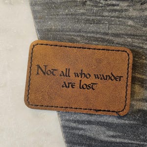May include: A rectangular, brown leather patch with the quote "Not all who wander are lost" in a dark font. The patch has a stitched black border and is set against a marbled background.