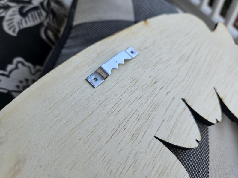 May include: Close-up of a wooden decorative piece with a metal sawtooth hanger attached. The wood has a light color and visible grain. The hanger is silver and has a series of teeth.