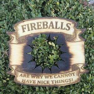 May include: Wooden sign with the words "FIREBALLS" and "ARE WHY WE CANNOT HAVE NICE THINGS!" in a banner design. The sign has a starburst cut-out in the center and is mounted on a green bush.
