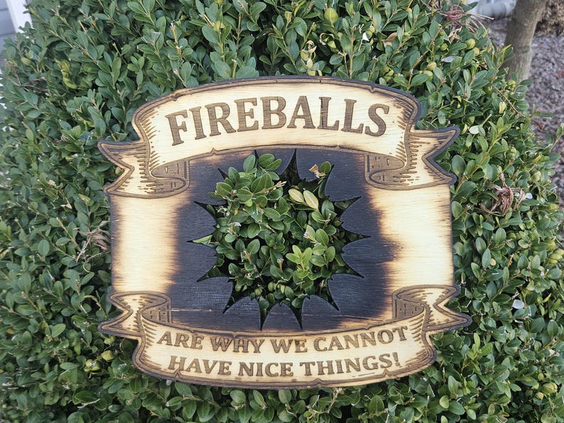 May include: Wooden sign with the words "FIREBALLS" and "ARE WHY WE CANNOT HAVE NICE THINGS!" in a banner design. The sign has a starburst cut-out in the center and is mounted on a green bush.