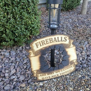 May include: A wooden sign with the word "FIREBALLS" in a banner design. The sign has a charred center with a hole and the text "ARE WHY WE CANNOT HAVE NICE THINGS!" The sign is outdoors near a bush and rocks.
