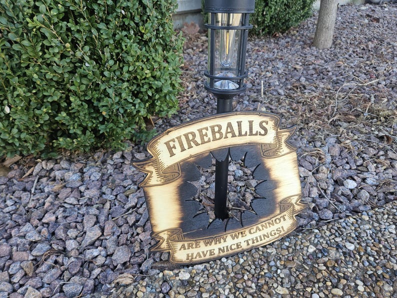 May include: A wooden sign with the word "FIREBALLS" in a banner design. The sign has a charred center with a hole and the text "ARE WHY WE CANNOT HAVE NICE THINGS!" The sign is outdoors near a bush and rocks.
