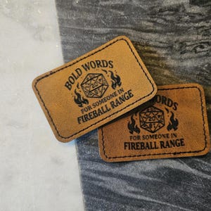 May include: Two rectangular brown leather patches with rounded corners. Each patch has the words "BOLD WORDS" at the top, a graphic of a 20-sided die, and the text "FOR SOMEONE IN FIREBALL RANGE" below. The patches are on a gray surface.