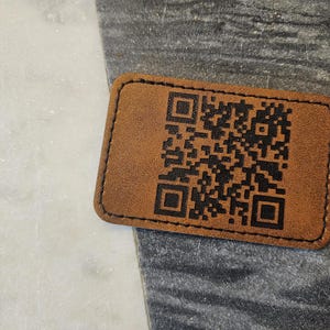 May include: A rectangular, brown leather patch with a black QR code printed on it. The patch has rounded corners and a black stitched border. The background is a mix of white and gray marble.