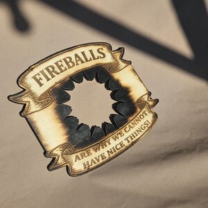 May include: Wooden coaster with a banner design. The banner reads "FIREBALLS" at the top and "ARE WHY WE CANNOT HAVE NICE THINGS!" at the bottom. The coaster has a circular cutout in the center and a burnt wood effect.