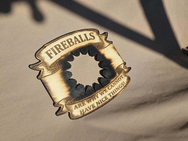 May include: Wooden coaster with a banner design. The banner reads "FIREBALLS" at the top and "ARE WHY WE CANNOT HAVE NICE THINGS!" at the bottom. The coaster has a circular cutout in the center and a burnt wood effect.