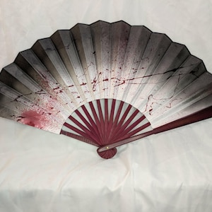 May include: A decorative folding fan with a dark grey and white design, accented with red splatters. The fan's base and ribs are a deep burgundy colour, creating a striking contrast. The fan is open, displaying its full, semi-circular shape.