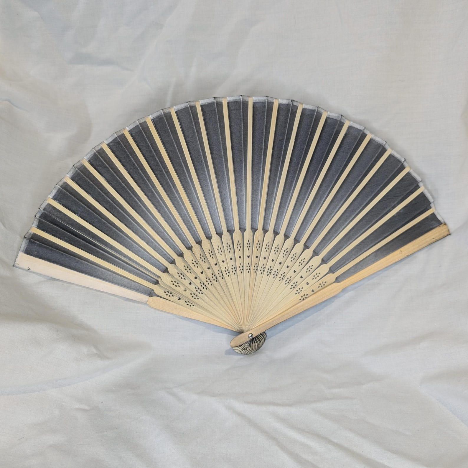 Folding hand fan with black ombré fabric and natural wood ribs, displayed on a light fabric background.