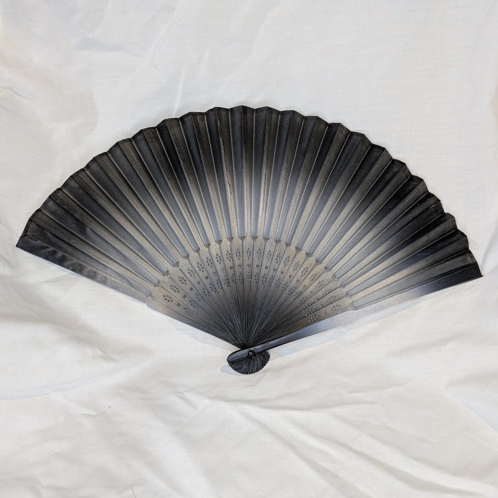 Black handheld folding fan with soft grey ombré fade, photographed on a white fabric background.