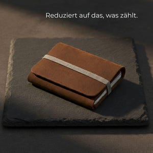 Könnte beinhalten: Ein braunes Lederportemonnaie mit einem grauen Gummibandverschluss. Das Portemonnaie ist rechteckig und scheint geschlossen zu sein. Der Text "Reduziert auf das, was zählt" ist im Bild zu sehen.