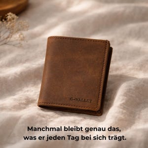 Könnte beinhalten: Ein braunes Lederportemonnaie mit dem Text "G-WALLET" wird auf einer weichen, strukturierten Oberfläche gezeigt. Das Portemonnaie ist rechteckig und scheint aus Leder mit Used-Look zu sein. Der Text "Manchmal bleibt genau das, was er jeden Tag bei sich trägt" steht unten.