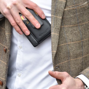 Op de afbeelding: Een zwarte leren portemonnee met de tekst G-WALLET in reli&euml;f op de voorzijde, voorzien van een kaartsleuf met een zichtbare kaart, gehouden tegen een wit overhemd onder een bruine en beige blazer met visgraatmotief en een subtiel ruitpatroon.