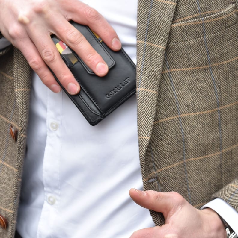Op de afbeelding: Een zwarte leren portemonnee met de tekst G-WALLET in reli&euml;f op de voorzijde, voorzien van een kaartsleuf met een zichtbare kaart, gehouden tegen een wit overhemd onder een bruine en beige blazer met visgraatmotief en een subtiel ruitpatroon.