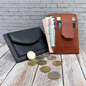 Op de afbeelding: Een zwarte leren portemonnee met klepsluiting en een bruine leren portemonnee met het opschrift G-WALLET en een verticale band, beide gevuld met papiergeld en kaarten, met verspreide gouden en zilveren munten op een houten ondergrond.