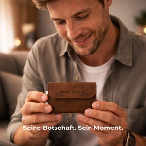Könnte beinhalten: Ein braunes Lederportemonnaie mit dem Text "Immer Dein" und einem Herzsymbol wird von einer Person gehalten. Das Portemonnaie hat einen Klappenverschluss. Der Text "Seine Botschaft. Sein Moment." steht unten im Bild.