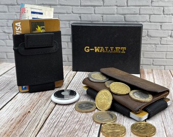 Op de afbeelding: Een zwarte elastische kaarthouder met een gouden creditcard en papiergeld, een bruine leren portemonnee met euro-munten, een witte ronde tracker en een zwarte doos met de gouden tekst G-WALLET op de voorzijde gedrukt.