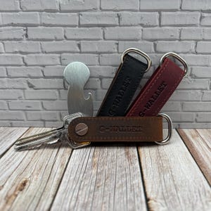 Op de afbeelding: Drie lederen sleutelorganizers in bruin, zwart en bordeauxrood, elk voorzien van de tekst G-WALLET. De bruine organizer bevat een zilverkleurige metalen sleutel en een zilverkleurige metalen flesopener bevindt zich achter de zwarte en bordeauxrode.